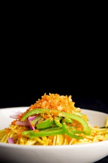 A close-up of a dish featuring a mound of spaghetti topped with vibrant green bell pepper slices and finely chopped purple onions. The dish is further garnished with a generous layer of golden, crispy breadcrumbs. The presentation is vivid against a stark black background, highlighting the colors and textures of the ingredients.