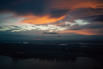 Vibrant orange and blue hues spread across the sky during sunset, with patches of dark clouds adding texture. Below, the silhouette of a cityscape is visible, with a river winding through the scene and some buildings faintly lit.