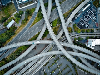 A complex network of highways and overpasses interweaves, forming a web of concrete. Vehicles can be seen traveling along the roads, surrounded by patches of greenery and urban infrastructure, including buildings and parking lots.