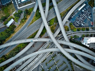 A complex network of highways and overpasses interweaves, forming a web of concrete. Vehicles can be seen traveling along the roads, surrounded by patches of greenery and urban infrastructure, including buildings and parking lots.