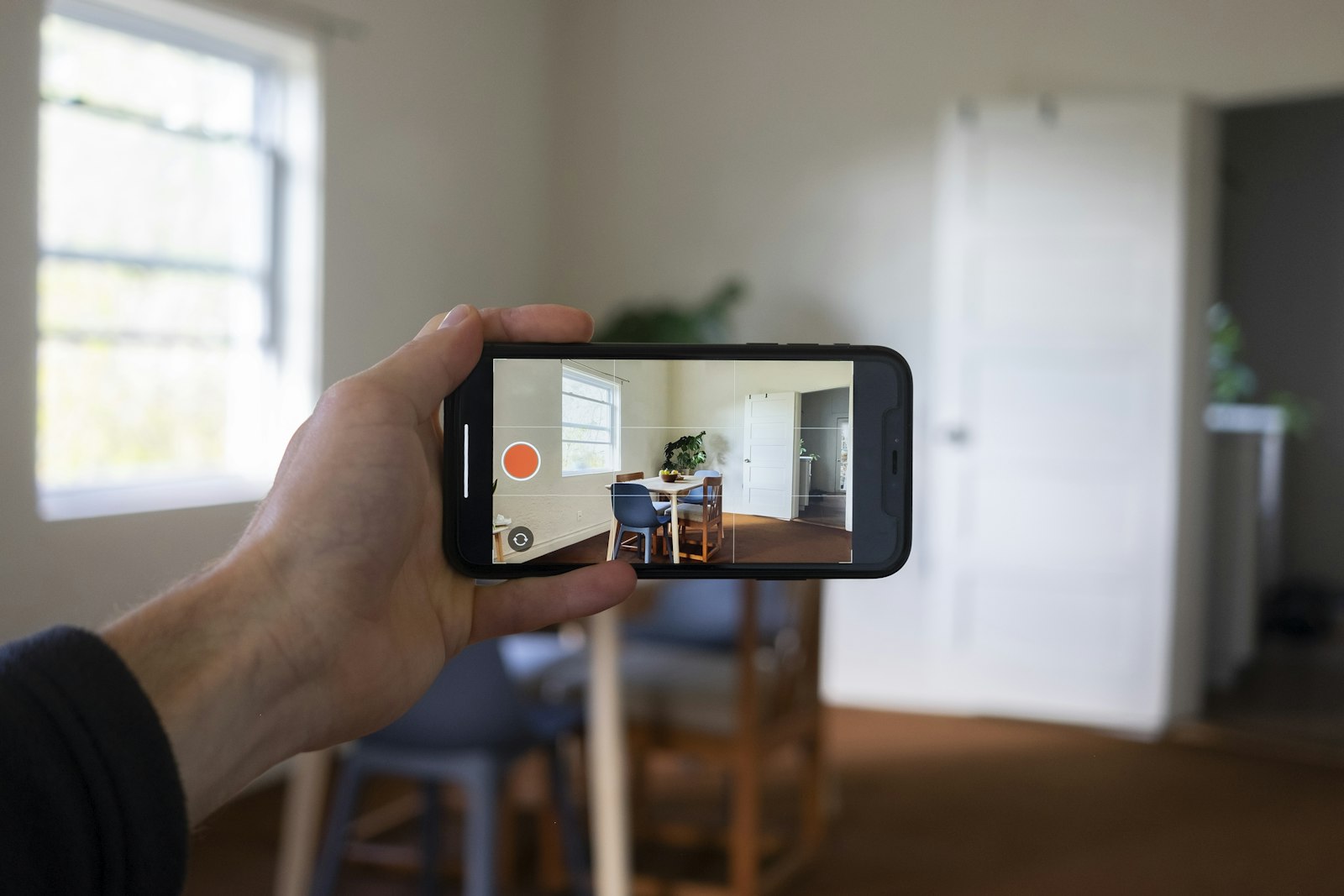 A hand holds up a smartphone recording an empty dining room through its camera, capturing the space on screen