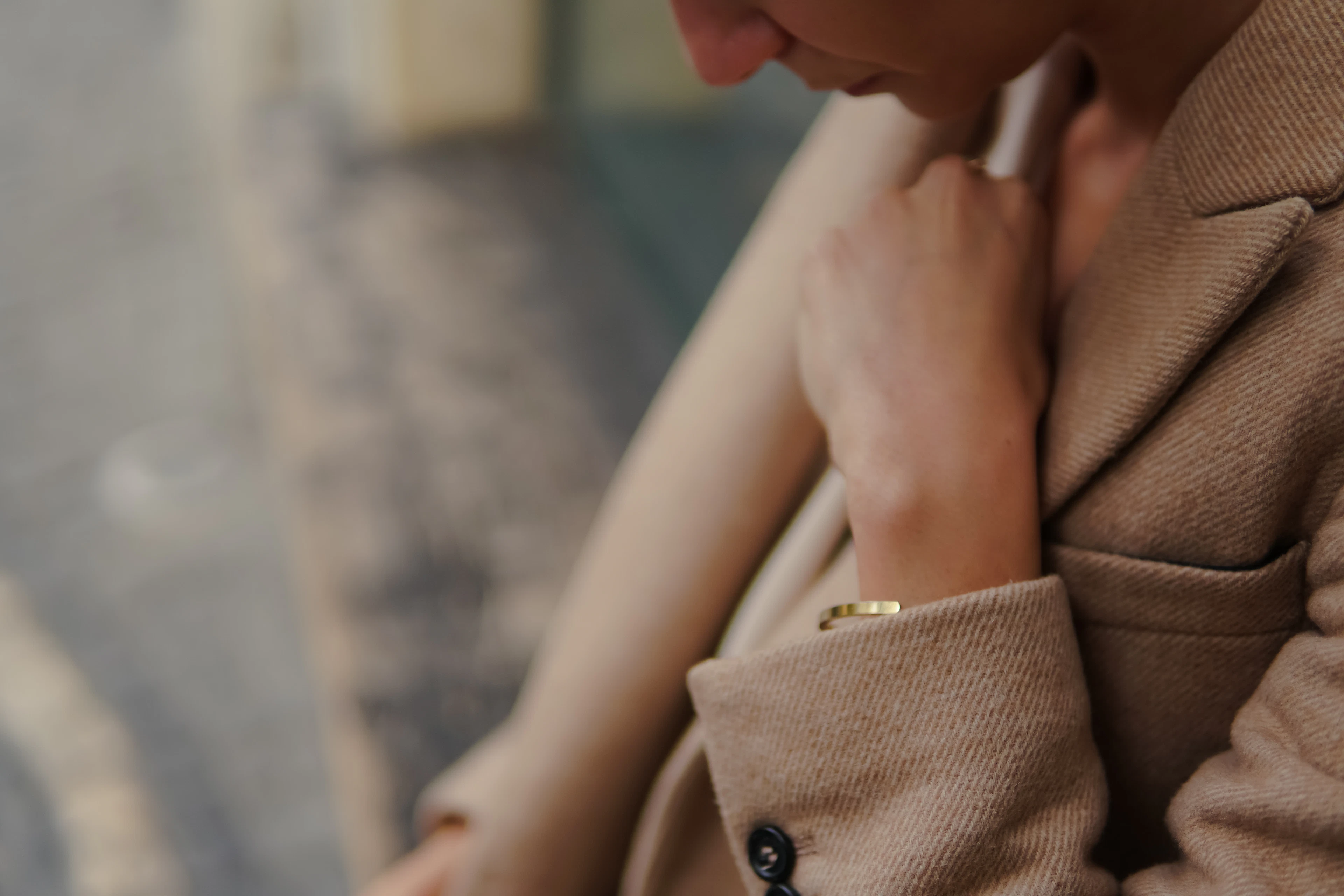 A hand with a fine gold bracelet resting on the lapel of a textured tan wool coat