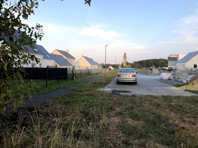 A suburban area with newly built houses on a quiet street. There is a lone car parked on the road, surrounded by grass and patches of dirt. In the background, a tall church tower is visible against the blue sky. A black metal fence runs along the left side.