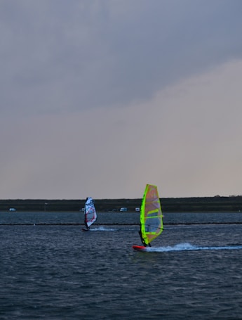 Two windsurfers glide across a large body of water under a cloudy sky. The water shows ripples created by the wind and movement of the boards. The sails are brightly colored, with one predominantly yellow and the other featuring red and blue hues. In the background, a strip of land stretches across the horizon with a few small structures visible.