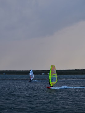 Two windsurfers glide across a large body of water under a cloudy sky. The water shows ripples created by the wind and movement of the boards. The sails are brightly colored, with one predominantly yellow and the other featuring red and blue hues. In the background, a strip of land stretches across the horizon with a few small structures visible.