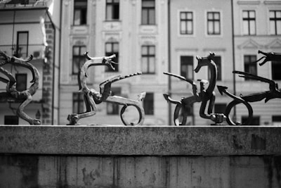 A series of abstract, whimsical sculptures depicting fantastical creatures with varying shapes and limbs, arranged on a stone ledge. The background consists of historical buildings with intricate architectural details and windows.
