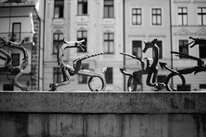 A series of abstract, whimsical sculptures depicting fantastical creatures with varying shapes and limbs, arranged on a stone ledge. The background consists of historical buildings with intricate architectural details and windows.