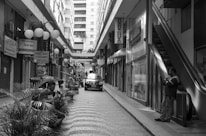 A monochrome street scene featuring a narrow pedestrian walkway lined with shops and closed storefronts. Several people are sitting and standing near potted plants along the path. An escalator is visible on the right side, with an individual standing near it. A small car is parked in the middle of the pathway, and various signs are hanging above or against the shop entrances on both sides, displaying text in Portuguese. The background features tall buildings, enhancing the urban setting.