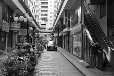 A monochrome street scene featuring a narrow pedestrian walkway lined with shops and closed storefronts. Several people are sitting and standing near potted plants along the path. An escalator is visible on the right side, with an individual standing near it. A small car is parked in the middle of the pathway, and various signs are hanging above or against the shop entrances on both sides, displaying text in Portuguese. The background features tall buildings, enhancing the urban setting.