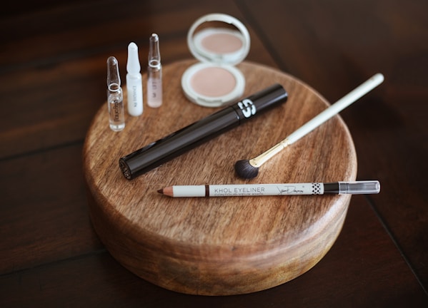 A collection of makeup items placed neatly on a round wooden platform, including small bottles of skincare products, a compact with powder, a tube of mascara, an eyeliner pencil, and a makeup brush. The arrangement is displayed on a polished dark wooden table.