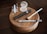 A collection of makeup items placed neatly on a round wooden platform, including small bottles of skincare products, a compact with powder, a tube of mascara, an eyeliner pencil, and a makeup brush. The arrangement is displayed on a polished dark wooden table.