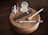 A collection of makeup items placed neatly on a round wooden platform, including small bottles of skincare products, a compact with powder, a tube of mascara, an eyeliner pencil, and a makeup brush. The arrangement is displayed on a polished dark wooden table.