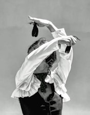 A person dressed in a traditional flamenco outfit with a ruffled shirt and a dark vest is performing a dance. The arms are elegantly raised above the head, holding castanets, which adds to the dynamic posture. The face is obscured by the raised arms, enhancing the focus on the expressive movement of the dance.