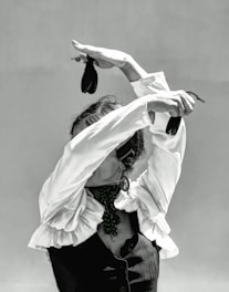 A person dressed in a traditional flamenco outfit with a ruffled shirt and a dark vest is performing a dance. The arms are elegantly raised above the head, holding castanets, which adds to the dynamic posture. The face is obscured by the raised arms, enhancing the focus on the expressive movement of the dance.