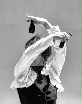A person dressed in a traditional flamenco outfit with a ruffled shirt and a dark vest is performing a dance. The arms are elegantly raised above the head, holding castanets, which adds to the dynamic posture. The face is obscured by the raised arms, enhancing the focus on the expressive movement of the dance.