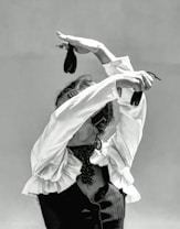 A person dressed in a traditional flamenco outfit with a ruffled shirt and a dark vest is performing a dance. The arms are elegantly raised above the head, holding castanets, which adds to the dynamic posture. The face is obscured by the raised arms, enhancing the focus on the expressive movement of the dance.