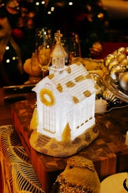 A small, decorative church-shaped ornament sits on a wooden block, illuminated from within. The church is adorned with glittery details and a wreath hanging at the front. Surrounding the ornament are festive elements including champagne glasses and shiny baubles. The background features a blurred Christmas tree with lights.