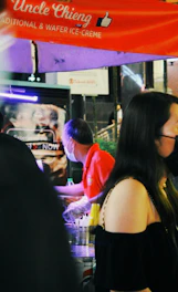 A street vendor wearing a red shirt and face mask prepares food at a stall under a canopy that reads 'Uncle Chieng Traditional & Wafer Ice-Crème'. People are gathered around, and there are various items on the counter, including containers and straws.