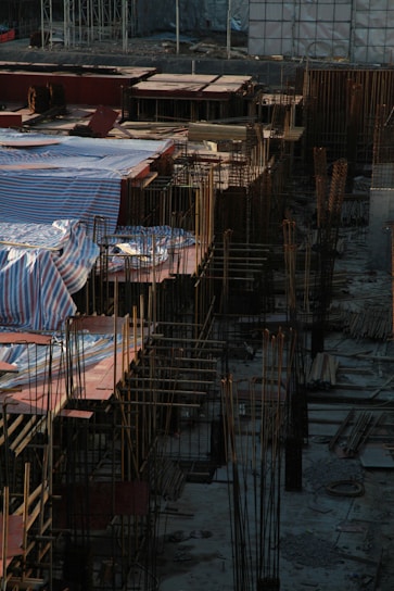 A construction site filled with steel rods and concrete foundations, with sections covered by striped tarpaulins. The scene captures the early stages of building, where rebar frameworks are prominently visible. The area is surrounded by various construction materials, including wooden planks and scaffolding.