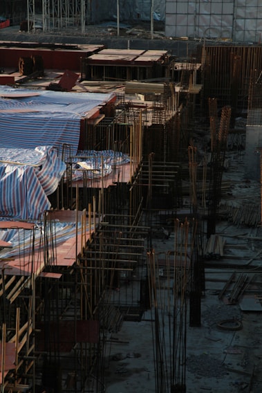 A construction site filled with steel rods and concrete foundations, with sections covered by striped tarpaulins. The scene captures the early stages of building, where rebar frameworks are prominently visible. The area is surrounded by various construction materials, including wooden planks and scaffolding.