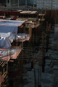 A construction site filled with steel rods and concrete foundations, with sections covered by striped tarpaulins. The scene captures the early stages of building, where rebar frameworks are prominently visible. The area is surrounded by various construction materials, including wooden planks and scaffolding.