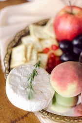 A platter featuring an assortment of foods, including a round of soft white cheese garnished with a sprig of rosemary, slices of soft yellow cheese, a red apple, a peach, clusters of red grapes, and other berries. The platter is placed on a wooden table.