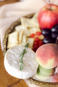 A platter featuring an assortment of foods, including a round of soft white cheese garnished with a sprig of rosemary, slices of soft yellow cheese, a red apple, a peach, clusters of red grapes, and other berries. The platter is placed on a wooden table.