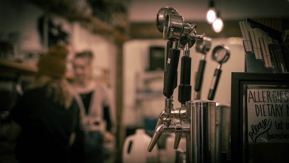 A close-up of coffee machine portafilters in focus, with a blurry background showing people, shelves, and light bulbs. A sign with dietary information is visible to the side.