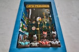 A decorative shop window framed by a blue border showcases four colorful ceramic busts of women wearing dotted clothing and headscarves. The window is filled with vibrant hanging decorations, including painted birds and hot air balloons, creating a lively and artistic atmosphere.