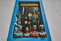 A decorative shop window framed by a blue border showcases four colorful ceramic busts of women wearing dotted clothing and headscarves. The window is filled with vibrant hanging decorations, including painted birds and hot air balloons, creating a lively and artistic atmosphere.