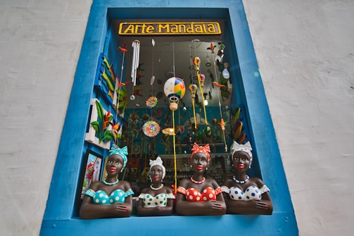A decorative shop window framed by a blue border showcases four colorful ceramic busts of women wearing dotted clothing and headscarves. The window is filled with vibrant hanging decorations, including painted birds and hot air balloons, creating a lively and artistic atmosphere.