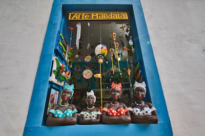 A decorative shop window framed by a blue border showcases four colorful ceramic busts of women wearing dotted clothing and headscarves. The window is filled with vibrant hanging decorations, including painted birds and hot air balloons, creating a lively and artistic atmosphere.