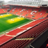 A large football stadium with vibrant red seats and the letters LFC prominently displayed across the seating. The field is well-maintained, with green grass, and sunlight or artificial lighting illuminating parts of it. A few people are scattered around the stadium, possibly staff or maintenance workers.