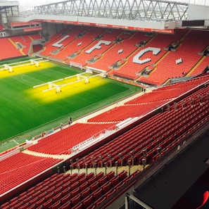 A large football stadium with vibrant red seats and the letters LFC prominently displayed across the seating. The field is well-maintained, with green grass, and sunlight or artificial lighting illuminating parts of it. A few people are scattered around the stadium, possibly staff or maintenance workers.