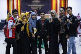 A group of eight young individuals is gathered together, some wearing matching outfits with red and white accents. One person holds a trophy, suggesting a celebratory event or competition setting. They stand in a row with a backdrop featuring festive orange and red drapes.