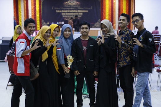 A group of eight young individuals is gathered together, some wearing matching outfits with red and white accents. One person holds a trophy, suggesting a celebratory event or competition setting. They stand in a row with a backdrop featuring festive orange and red drapes.