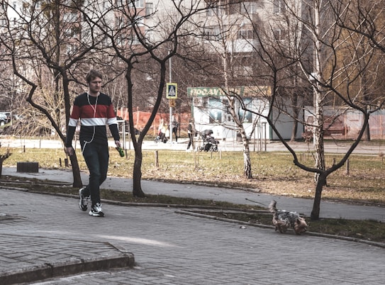 A person walking on a sidewalk under leafless trees, listening to music with earphones. They are wearing a sweater with horizontal stripes and holding a leash attached to a small dog. In the background, several people, including a person pushing a stroller, can be seen near a building.