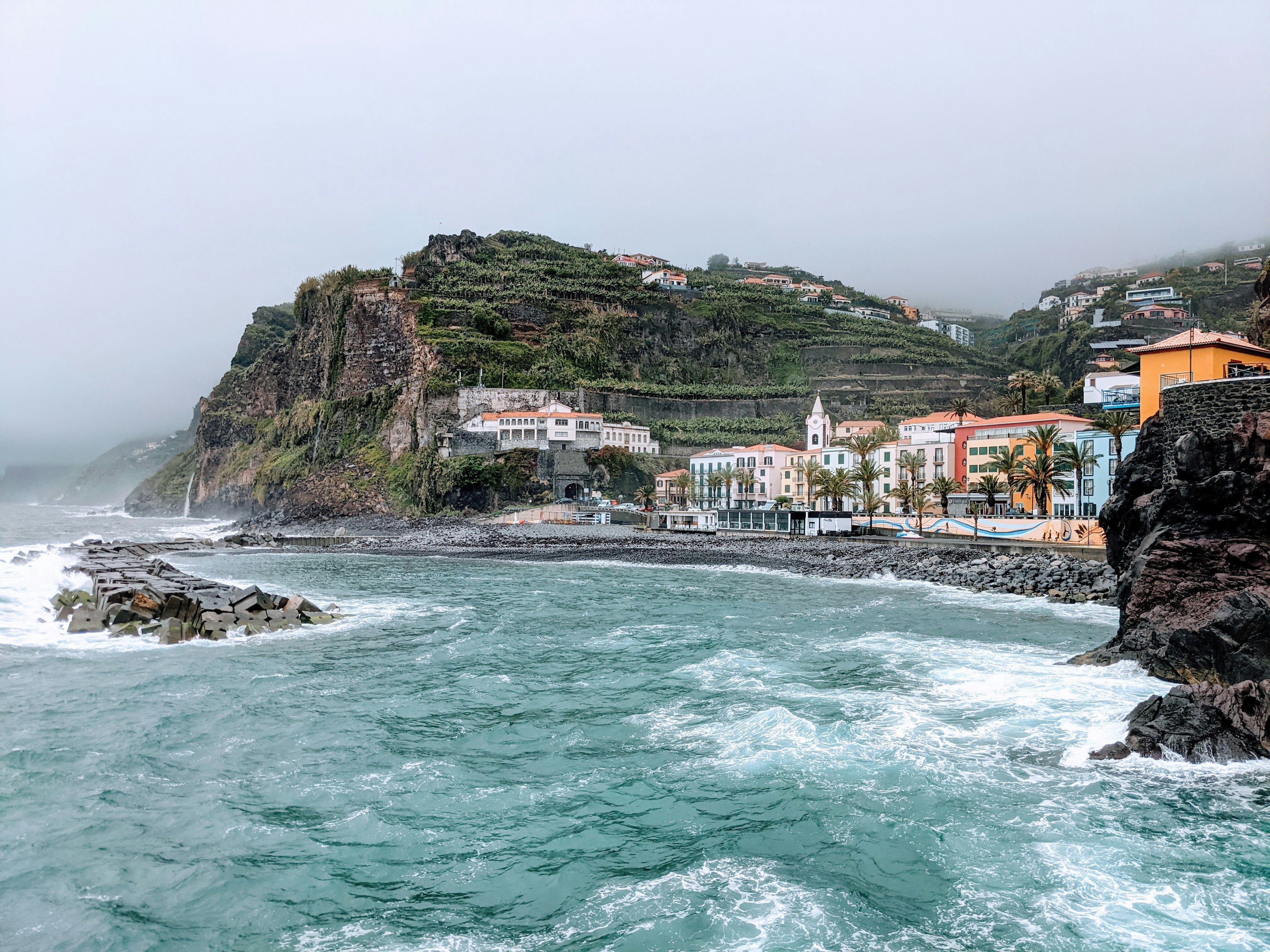 a body of water with houses on a hill in the background, Ponta do Sol in Madeira welcomes you with colorful facades of various buildings in the small village in the middle of the South coast of Madeira. This place looks magical: during storm, sunny weather or middle of the night.