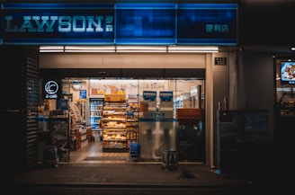 An illuminated storefront of a convenience store with a blue sign displaying 'LAWSON'. The entrance reveals a well-organized interior with shelves stocked with various grocery items and snacks. Posters and advertisements are visible on the glass doors, and the warm lighting creates a welcoming atmosphere.