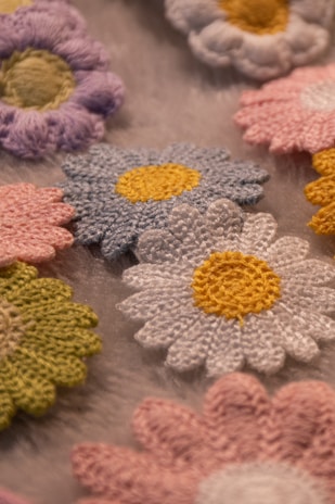 Various crocheted flowers are arranged on a soft, textured background. The flowers feature different colors, including white, pink, blue, yellow, and green, each with a distinct yellow or green center.