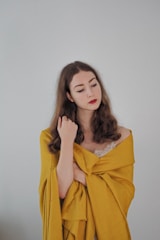 A woman with long brown hair wrapped in a yellow fabric, with a calm and introspective expression. She is wearing a lace top underneath, and her eyes are closed.