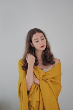 A woman with long brown hair wrapped in a yellow fabric, with a calm and introspective expression. She is wearing a lace top underneath, and her eyes are closed.