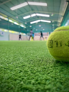 A close-up view of a tennis ball on a green indoor court with blurred figures in the background playing. The court has an overhead roof with white fluorescent lights.