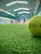A close-up view of a tennis ball on a green indoor court with blurred figures in the background playing. The court has an overhead roof with white fluorescent lights.