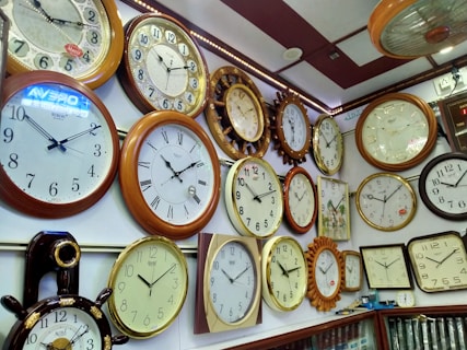 A collection of various wall clocks displayed on a shop wall. The clocks have different styles, sizes, and shapes, with most having white or cream faces and black numerals. Some clocks feature Roman numerals while others have standard numbering. The frames are mostly in shades of brown, gold, and wood textures, with a few digital elements visible in the background.