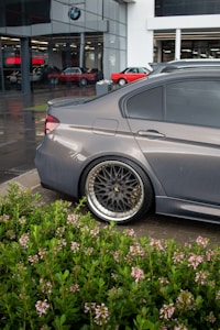 A sleek, gray car with aftermarket rims is parked in an outdoor setting, possibly near a dealership indicated by the building in the background. The foreground showcases lush green plants with small pink flowers, contrasting with the modern architectural features and other vintage cars visible through the glass.