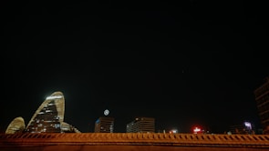 A night scene of a cityscape features several illuminated skyscrapers. One of the notable buildings displays a logo with the letters KPMG, while another building shows a Mercedes-Benz emblem. The architecture is highlighted against a deep, dark sky with minimal lighting elsewhere in the frame.