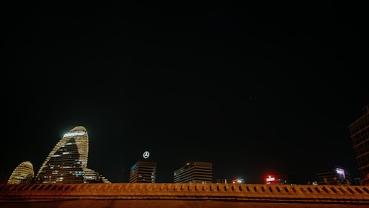 A night scene of a cityscape features several illuminated skyscrapers. One of the notable buildings displays a logo with the letters KPMG, while another building shows a Mercedes-Benz emblem. The architecture is highlighted against a deep, dark sky with minimal lighting elsewhere in the frame.