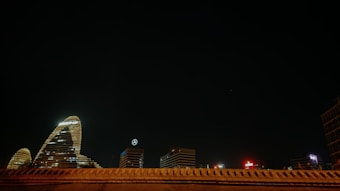 A night scene of a cityscape features several illuminated skyscrapers. One of the notable buildings displays a logo with the letters KPMG, while another building shows a Mercedes-Benz emblem. The architecture is highlighted against a deep, dark sky with minimal lighting elsewhere in the frame.