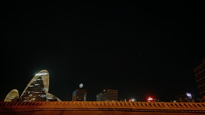 A night scene of a cityscape features several illuminated skyscrapers. One of the notable buildings displays a logo with the letters KPMG, while another building shows a Mercedes-Benz emblem. The architecture is highlighted against a deep, dark sky with minimal lighting elsewhere in the frame.